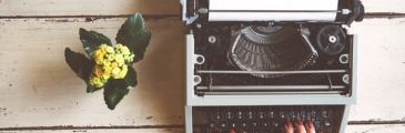 Mains de femme devant une machine à écrire avec un petit pot de fleurs jaunes sur la gauche
