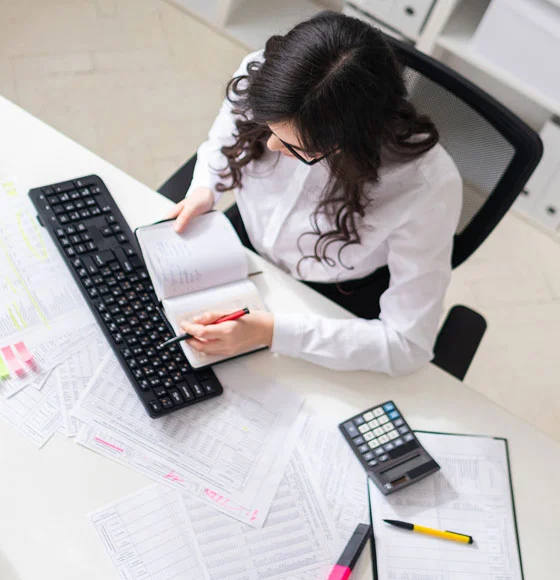 Une jeune fille est assise à un ordinateur du bureau, un stylo à la main et regardant le bloc-notes.